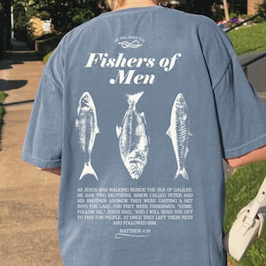 May include: Blue t-shirt with the text "Fishers of Men" and three fish illustrations. The back of the shirt also includes a Bible verse from Matthew 4:19. The shirt is a relaxed fit.