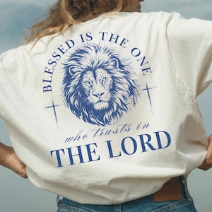 Może przedstawiać: Biała koszulka z niebieską grafiką lwa i napisem "BLESSED IS THE ONE who trusts in THE LORD." Koszulka ma krótkie rękawy i luźny krój. Słowo "White" znajduje się w lewym górnym rogu.
