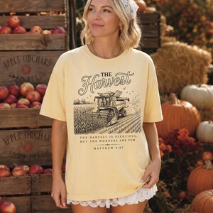 May include: A pale yellow short-sleeve t-shirt featuring a black graphic of a combine harvester in a field. The text reads: THE HARVEST. THE HARVEST IS PLENTIFUL, BUT THE WORKERS ARE FEW. MATTHEW 9:37. The shirt has a white lace trim along the bottom hem.
