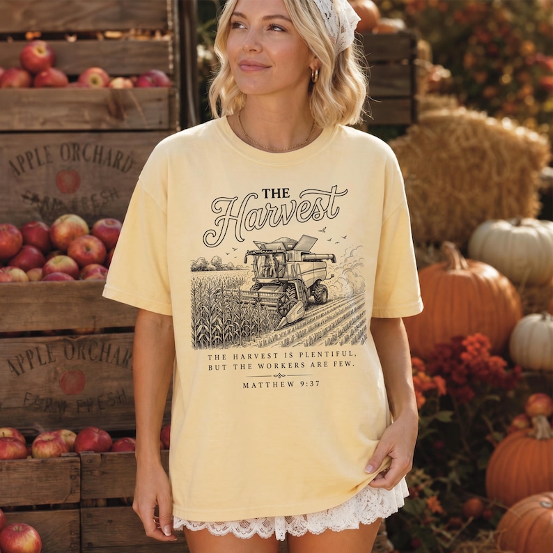 May include: A pale yellow short-sleeve t-shirt featuring a black graphic of a combine harvester in a field. The text reads: THE HARVEST. THE HARVEST IS PLENTIFUL, BUT THE WORKERS ARE FEW. MATTHEW 9:37. The shirt has a white lace trim along the bottom hem.
