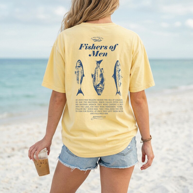May include: A pale yellow t-shirt featuring the words "Fishers of Men" and three blue fish designs. The back of the shirt also includes a Bible verse from Matthew 4:19. The shirt is a relaxed fit.