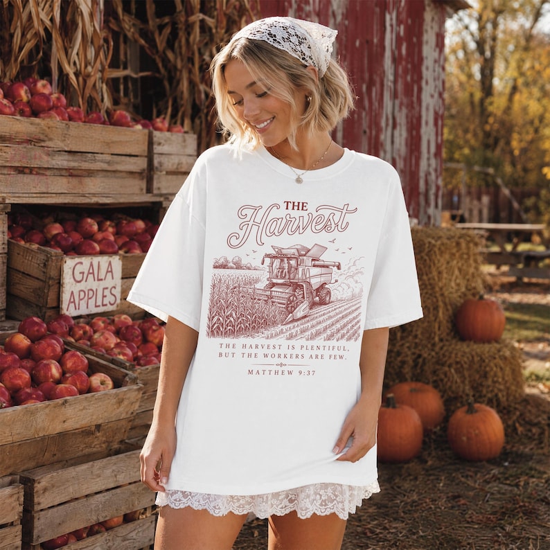May include: A white short-sleeved t-shirt featuring a maroon graphic of a combine harvester in a field. The text reads THE HARVEST, THE HARVEST IS PLENTIFUL, BUT THE WORKERS ARE FEW, and MATTHEW 9:37. The shirt has a white lace trim along the bottom hem.