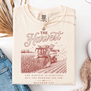May include: A cream-colored t-shirt featuring a maroon graphic of a combine harvester in a cornfield. The text reads THE HARVEST, followed by THE HARVEST IS PLENTIFUL, BUT THE WORKERS ARE FEW, and MATTHEW 9:37. The shirt has a Comfort Colors brand label.