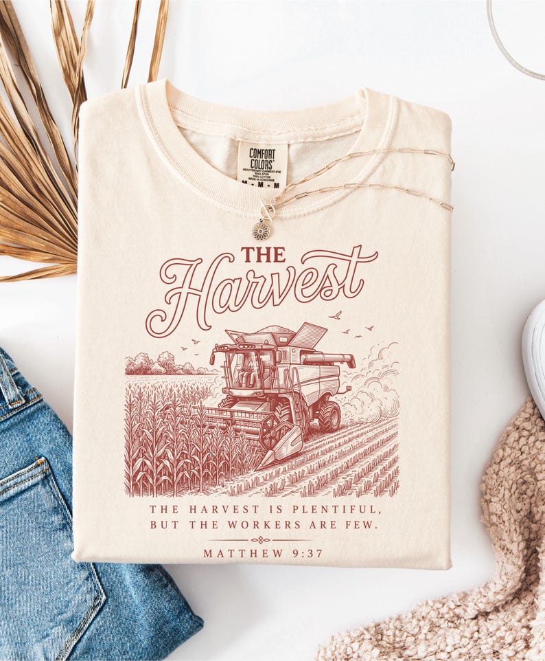May include: A cream-colored t-shirt featuring a maroon graphic of a combine harvester in a cornfield. The text reads THE HARVEST, followed by THE HARVEST IS PLENTIFUL, BUT THE WORKERS ARE FEW, and MATTHEW 9:37. The shirt has a Comfort Colors brand label.