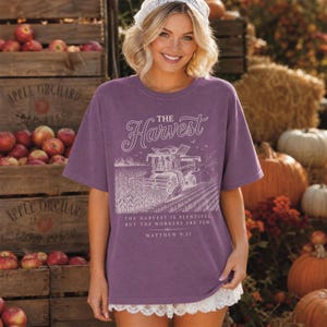 May include: A purple short-sleeve t-shirt featuring a white graphic of a combine harvester in a field. The text reads: The Harvest. The harvest is plentiful, but the workers are few. Matthew 9:37. The shirt has a crew neckline and a loose fit.