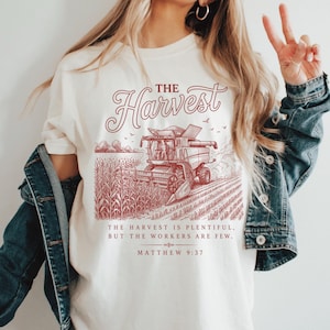 May include: A cream-colored t-shirt featuring a red graphic of a combine harvester in a cornfield. The text reads THE HARVEST, THE HARVEST IS PLENTIFUL, BUT THE WORKERS ARE FEW, and MATTHEW 9:37. The shirt is worn under a blue denim jacket.