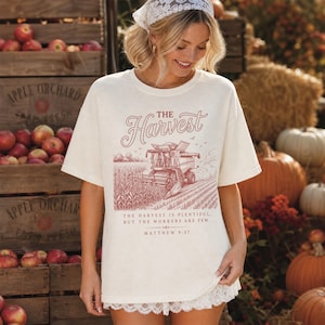 May include: An off-white short-sleeve t-shirt featuring a red graphic of a combine harvester in a cornfield. The text reads THE HARVEST, THE HARVEST IS PLENTIFUL, BUT THE WORKERS ARE FEW, and MATTHEW 9:37. The shirt has a crew neckline and a relaxed fit.