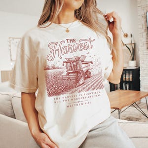 May include: A cream-colored t-shirt featuring a dark red graphic of a combine harvester in a cornfield. The text reads THE HARVEST, followed by THE HARVEST IS PLENTIFUL, BUT THE WORKERS ARE FEW. MATTHEW 9:37 in a decorative serif and script font.