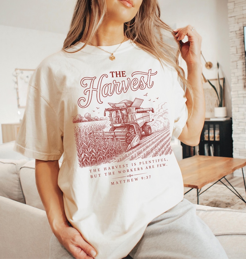 May include: A cream-colored t-shirt featuring a dark red graphic of a combine harvester in a cornfield. The text reads THE HARVEST, followed by THE HARVEST IS PLENTIFUL, BUT THE WORKERS ARE FEW. MATTHEW 9:37 in a decorative serif and script font.