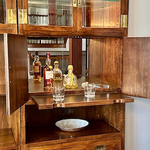 May include: A wooden bar cabinet with glass doors and brass hardware. Inside, various liquor bottles, glassware, and a decorative bowl are displayed. The cabinet is open, revealing a mirrored back and a pull-out shelf.