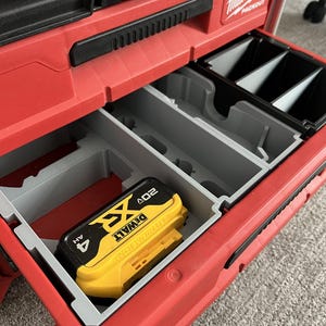 May include: A red Milwaukee Packout tool storage unit with an open drawer containing grey plastic dividers and a yellow and black DeWalt 20V XR 4AH lithium-ion battery. The storage unit features a black handle and the Milwaukee Packout logo.