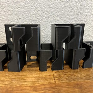 May include: A set of six black 3D-printed plastic mounting brackets, each featuring a square-shaped frame with a notched side profile and a single circular screw hole on the flat mounting tab for secure attachment to a flat surface.