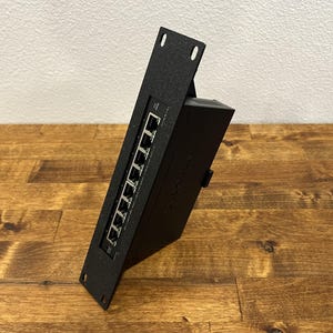 May include: A black metal rack-mount network switch featuring eight RJ45 Ethernet ports arranged vertically on the front panel. The device includes mounting flanges with screw holes at the top and bottom, and a textured finish on the exterior.