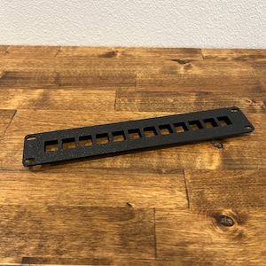 May include: A black rectangular rack mount panel featuring a series of twelve evenly spaced square cutouts along the center and four circular mounting holes located at each of the four corners of the flat, textured metal component.