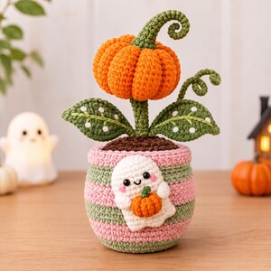May include: A crocheted orange pumpkin with a green curly stem and two green leaves with white dots, planted in a striped pink and green pot. The pot features a white ghost appliqué holding a small orange pumpkin on its front surface.
