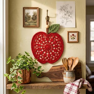 Puede incluir: Un adorno de pared con forma de manzana tejida a ganchillo en color rojo con una hoja verde, colgado de un cordón marrón con una cuenta de madera en un gancho de metal. La manzana presenta un intrincado patrón de encaje circular en su cuerpo.