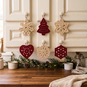 May include: Six crocheted ornaments in cream and deep red colors, featuring shapes of a snowflake, a Christmas tree, a star, a heart, a circular doily, and a hexagon, each hanging from a brown cord on white cabinet knobs.
