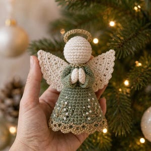 May include: A crocheted angel ornament featuring a cream-colored head and wings, a sage green dress with white bead accents, and a gold metallic halo and hem. The angel has hands clasped in front and is crafted with detailed yarn stitching throughout.