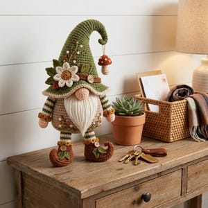 Pode incluir: Uma figura de gnomo em crochê com um chapéu alto verde-oliva adornado com uma flor branca e um pequeno pingente de cogumelo pendurado. O gnomo tem um corpo bege, uma longa barba de fio branco e sapatos castanhos curvados com folhas.