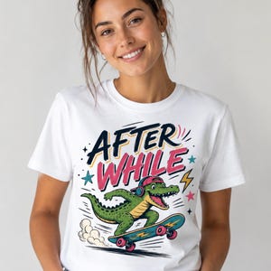 May include: A white short-sleeve t-shirt featuring a graphic of a green crocodile wearing a pink cap and headphones while riding a blue skateboard. The text "AFTER WHILE" is printed above the crocodile in bold black and pink lettering.