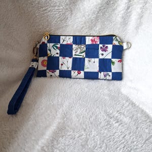 Op de afbeelding: Een rechthoekig polstasje met een blauw-wit geblokt patroon en bloemenprints op de witte vierkanten. Het tasje is voorzien van een goudkleurige rits met een hartvormige trekker en een bijpassende blauwe stoffen polsband aan de zijkant.