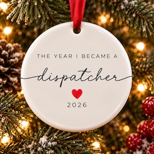 May include: A round white ceramic ornament hanging from a red ribbon. The text reads THE YEAR I BECAME A dispatcher with a small red heart icon below the word and the year 2026 printed at the bottom in a simple black sans-serif font.