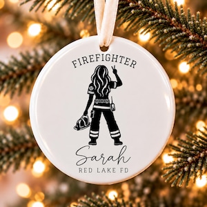 Peut inclure: Une décoration ronde en céramique blanche avec un motif noir représentant un pompier aux cheveux longs tenant un casque et faisant un signe de paix. Le texte indique FIREFIGHTER, Sarah et RED LAKE FD. Un ruban crème est fixé au sommet.