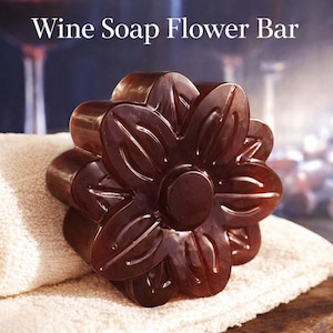 Könnte beinhalten: Ein dunkelbraunes, blütenförmiges Seifenstück mit dem weißen Text "Wine Soap Flower Bar". Die Seife liegt auf einem weißen Handtuch. Die Seife hat ein Blumenmuster und ist wahrscheinlich nach Wein duftend.