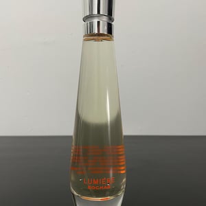 Puede incluir: Un frasco de perfume de vidrio transparente con tapa plateada. El frasco tiene forma cónica y contiene un líquido de color claro. El texto "LUMIERE ROCHAS" está impreso en naranja cerca de la base. El frasco está sobre una superficie oscura.