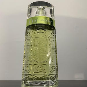Può includere: Un flacone di profumo rettangolare con un corpo in vetro trasparente strutturato e un tappo trasparente. Il flacone contiene un liquido verde chiaro. L'etichetta recita "O de Lancôme" in verde e bianco. Il flacone ha un vaporizzatore color argento.