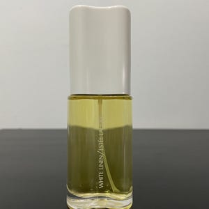 Peut inclure: Flacon de parfum en verre transparent avec un bouchon blanc. Le flacon contient un liquide jaune et porte l'inscription verticale "WHITE LINEN / ESTEE LAUDER". Le flacon est posé sur une surface sombre.