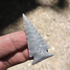 May include: A light gray, triangular stone arrowhead featuring a sharp, pointed tip and a notched base. The surface of the object displays a textured, flaked pattern characteristic of knapped stone, with distinct ridges along the edges.