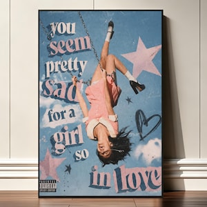 Puede incluir: Un póster rectangular que muestra a una persona colgada boca abajo de un columpio sobre un fondo de cielo azul. El texto dice, you seem pretty sad for a girl so in love. El diseño incluye estrellas rosas, un corazón y un marco negro.