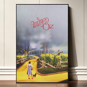 May include: A framed vertical poster featuring The Wizard of Oz with red text at the top. The scene depicts a person in a blue dress and a small dog walking along a yellow brick road through a cornfield, with a scarecrow standing nearby.