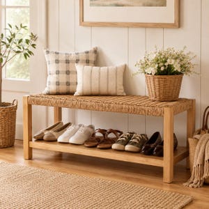 May include: A wooden entryway bench with a woven seat and a lower shelf for shoe storage. The bench is topped with a square plaid pillow and a rectangular striped pillow, alongside a woven basket containing white flowers and greenery.