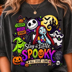Puede incluir: Una camiseta gris carbón con un estampado gráfico colorido de Jack Skellington, Sally y Zero de Pesadilla antes de Navidad. El diseño incluye calabazas, murciélagos, una casa encantada y el texto Stay a Little SPOOKY ALL YEAR LONG.