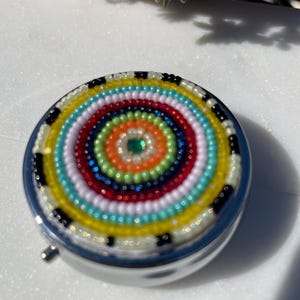 May include: A round, silver pill box with a vibrant, beaded design. The beaded pattern features concentric circles of various colors, including red, yellow, blue, and white. A small, green gem is in the center.
