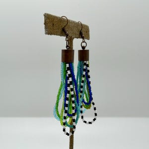 May include: Beaded earrings with copper-colored metal accents. The earrings feature multiple strands of colorful beads in shades of green, blue, black, and white. The earrings are displayed on a brown earring stand.