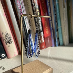 May include: A pair of blue, white, and black beaded dangle earrings hanging on a wooden stand. The earrings are made of small beads and have a silver hook.