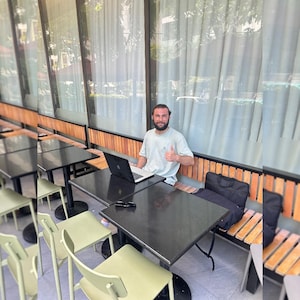 Op de afbeelding: Een persoon met een baard draagt een lichtgekleurd t-shirt en zit aan een zwarte vierkante tafel met een laptop. De persoon geeft een duim omhoog. De setting bevat lichtgroene stoelen en een houten bank tegen een raamwand.