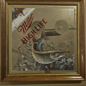 May include: A rectangular mirror in a brown wooden frame featuring a Miller High Life logo in red text. The mirror displays an illustration of a fish swimming underwater, a winter scene with a person walking on ice, and the text TIP-UP in the corner.
