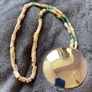 May include: A necklace featuring a large, polished gold-tone pendant. The necklace has a strand of rectangular, off-white beads, interspersed with green beads. The pendant reflects the surrounding environment.