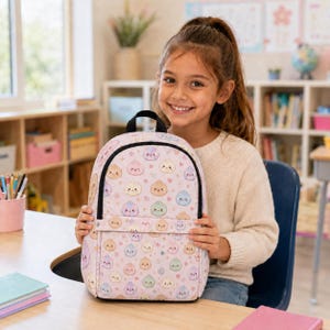 Könnte beinhalten: Ein hellrosa Rucksack mit einem durchgehenden Muster aus kleinen, bunten, lächelnden Knödel-Figuren in Blau-, Grün-, Gelb- und Violetttönen, akzentuiert durch winzige Herzen und Sterne, mit einem schwarzen Tragegriff und schwarzem Reißverschluss.