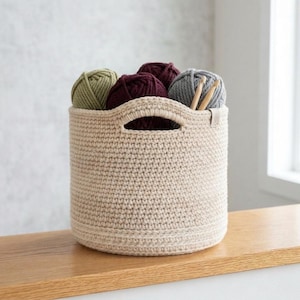 May include: A beige crochet basket filled with yarn balls in shades of green, burgundy, and gray. Two wooden knitting needles are visible. The basket has two handles and sits on a wooden surface.