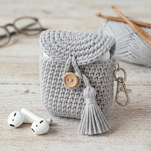 May include: A gray crocheted AirPods case with a wooden button closure and a matching tassel. The case has a silver-colored clasp and is next to a pair of white wireless earbuds. A ball of yarn and crochet hooks are in the background.