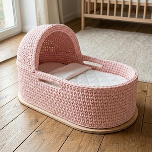 May include: A pink crocheted baby bassinet with a rounded hood and handles. The bassinet sits on a wooden base and contains a white quilted mattress and a small pillow and blanket. The bassinet is designed for a baby.