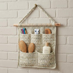 May include: A crocheted wall organizer in cream and beige, featuring multiple pockets for storage. It hangs from a wooden dowel and a braided cord. The pockets hold items like brushes, pens, a comb, and lotion.