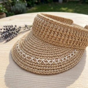 May include: A beige crocheted sun visor featuring a wide, structured brim and a tall, textured headband. The brim is decorated with a horizontal row of white cross-stitch embroidery detailing that runs along the front edge of the visor.