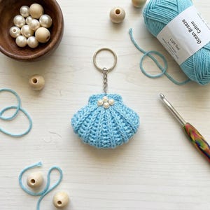 May include: A light blue, crocheted seashell keychain with three pearl accents. The keychain is attached to a silver ring. The image also shows a ball of light teal yarn labeled "Ocean Breeze Cotton", wooden beads, and a crochet hook.