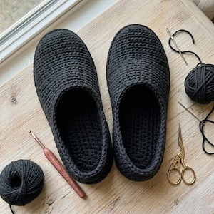 Puede incluir: Un par de zapatillas negras de ganchillo, con un ovillo de hilo a juego, un ganchillo, una aguja y unas tijeras. Las zapatillas están hechas a mano y parecen suaves y cómodas. Los artículos están dispuestos sobre una superficie de madera clara.